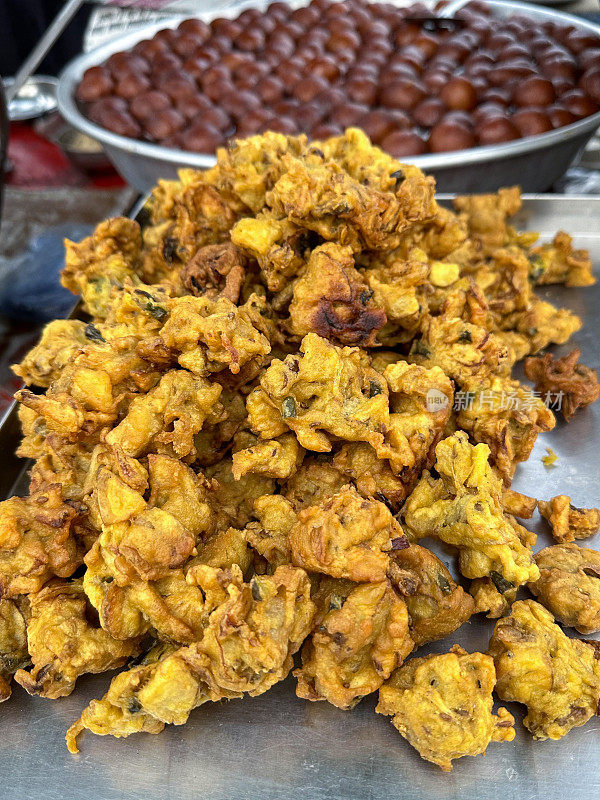 特写图片:矩形餐盘上的一堆酥脆的洋葱bhajis，印度街头美食素食小吃，背景是不锈钢碗的Gulab jamun(玫瑰水浆果)Mithai(糖果)，高视图，重点在前景